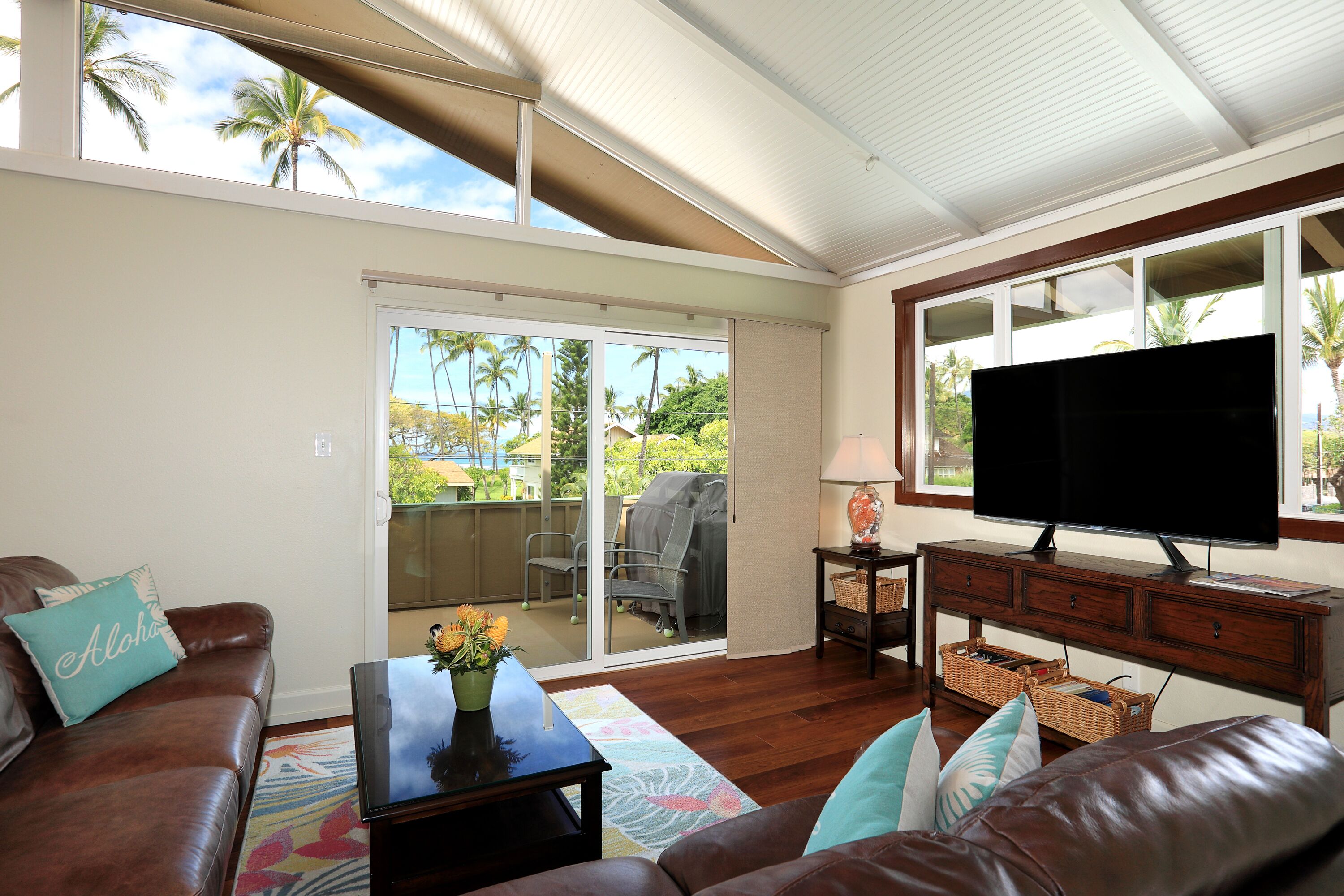 Un salón con un sofá de cuero marrón, una mesa de centro con un florero, un televisor de pantalla plana sobre un mueble de madera y un ventanal corredero que da a un patio exterior con vistas a palmeras.
