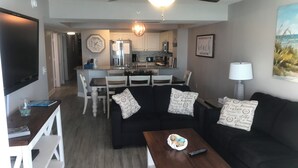 Ein Wohnzimmer mit einer schwarzen Couch, einem Couchtisch und einem Fernseher. Im Hintergrund ist eine Küche mit weißen Schränken und einem Essbereich zu sehen.