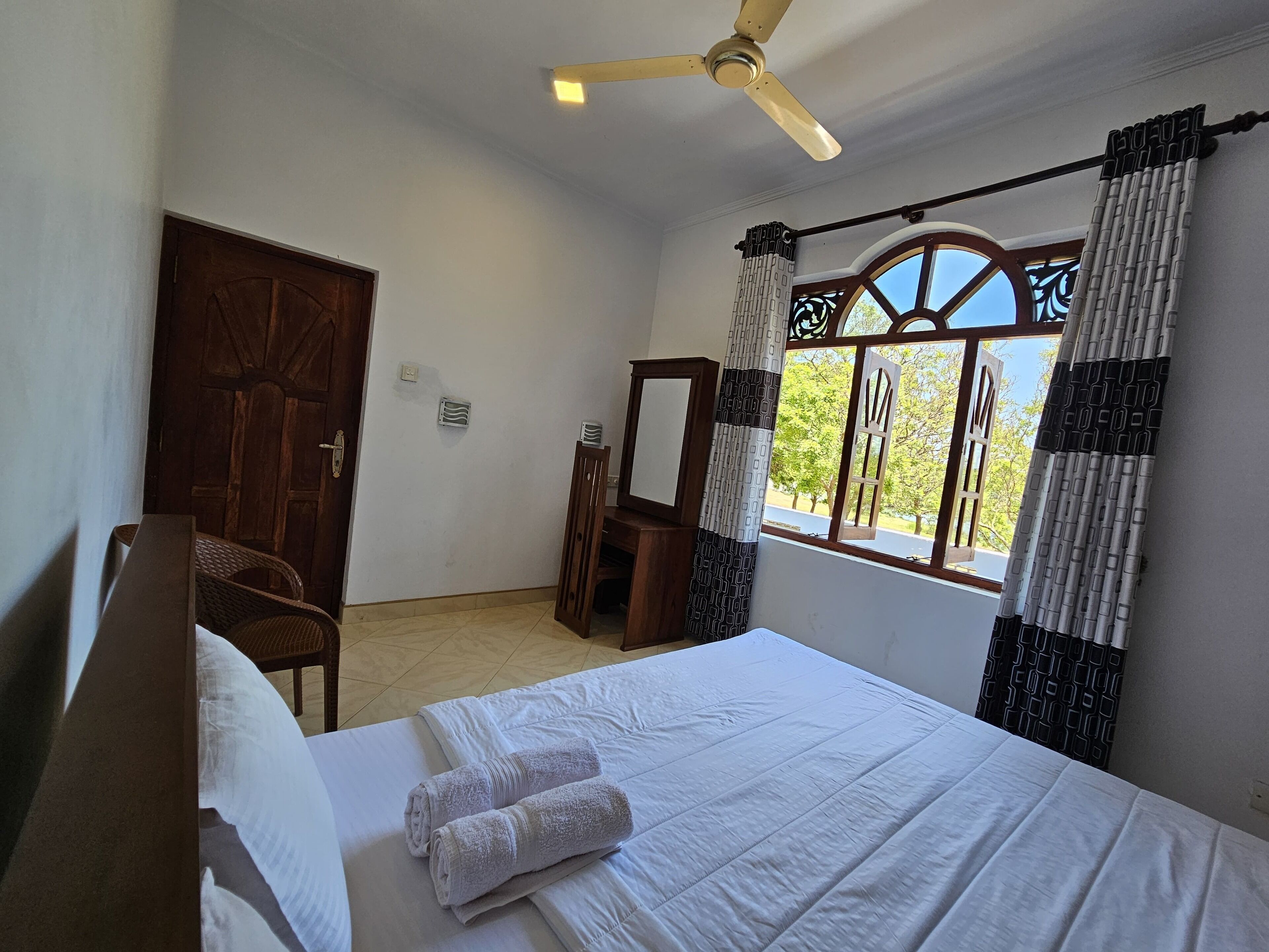 A hotel room with a bed, a chair, a ceiling fan, a window with curtains, and a door.