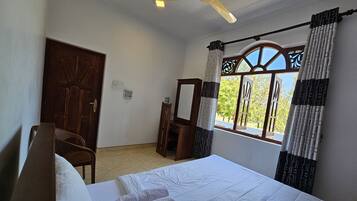 A hotel room with a bed, a chair, a ceiling fan, a window with curtains, and a door.