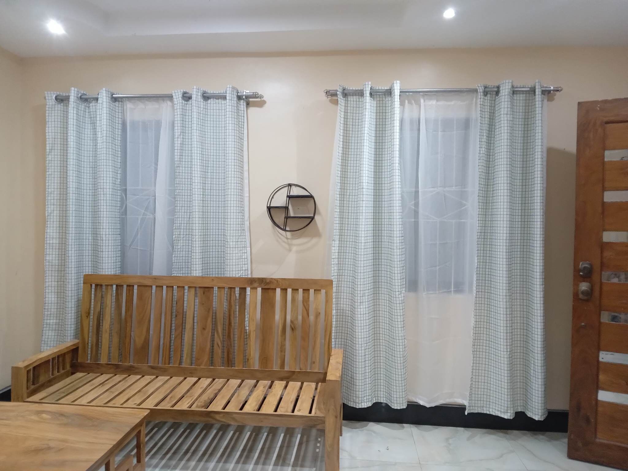 Una habitación con un banco de madera, dos ventanas con cortinas a cuadros, un estante redondo en la pared y una puerta de madera.