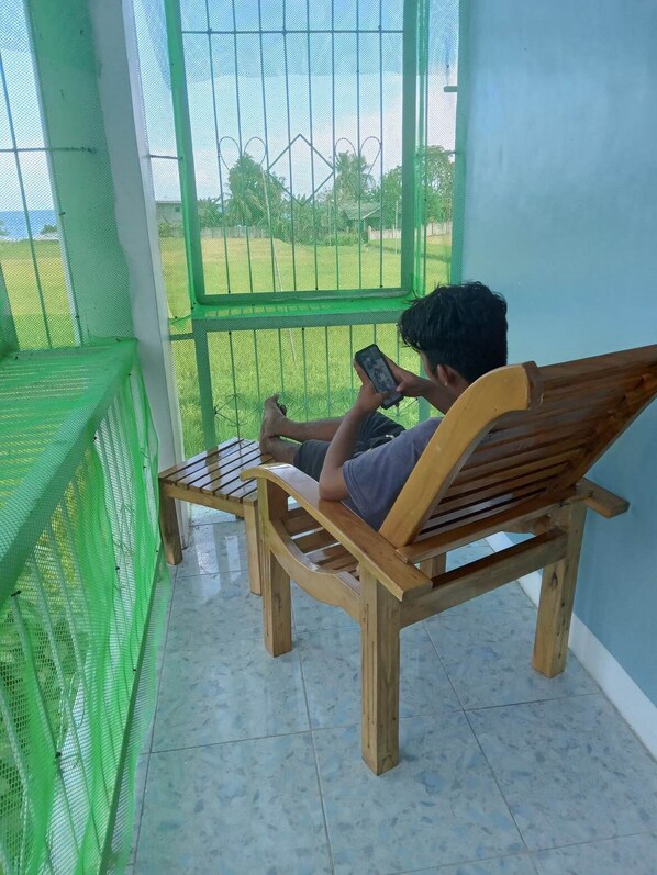 A person sitting on a wooden bench in a covered outdoor area with a green mesh fence, reading a book.