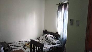 A small, sparsely furnished room with a single bed, a wooden chair, a desk with a chair, and a window with curtains.