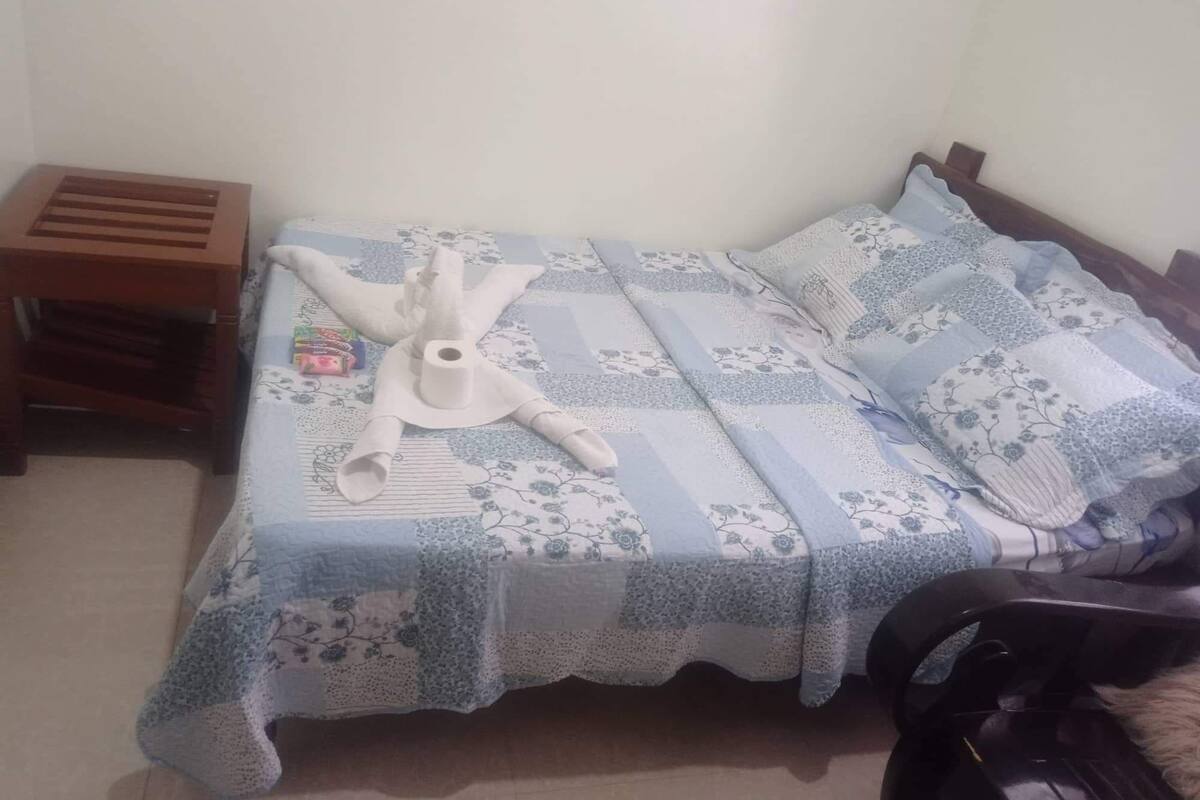 Un lit recouvert d’une couvre-lit à motifs floraux, une table de chevet en bois et un objet blanc posé sur le lit.