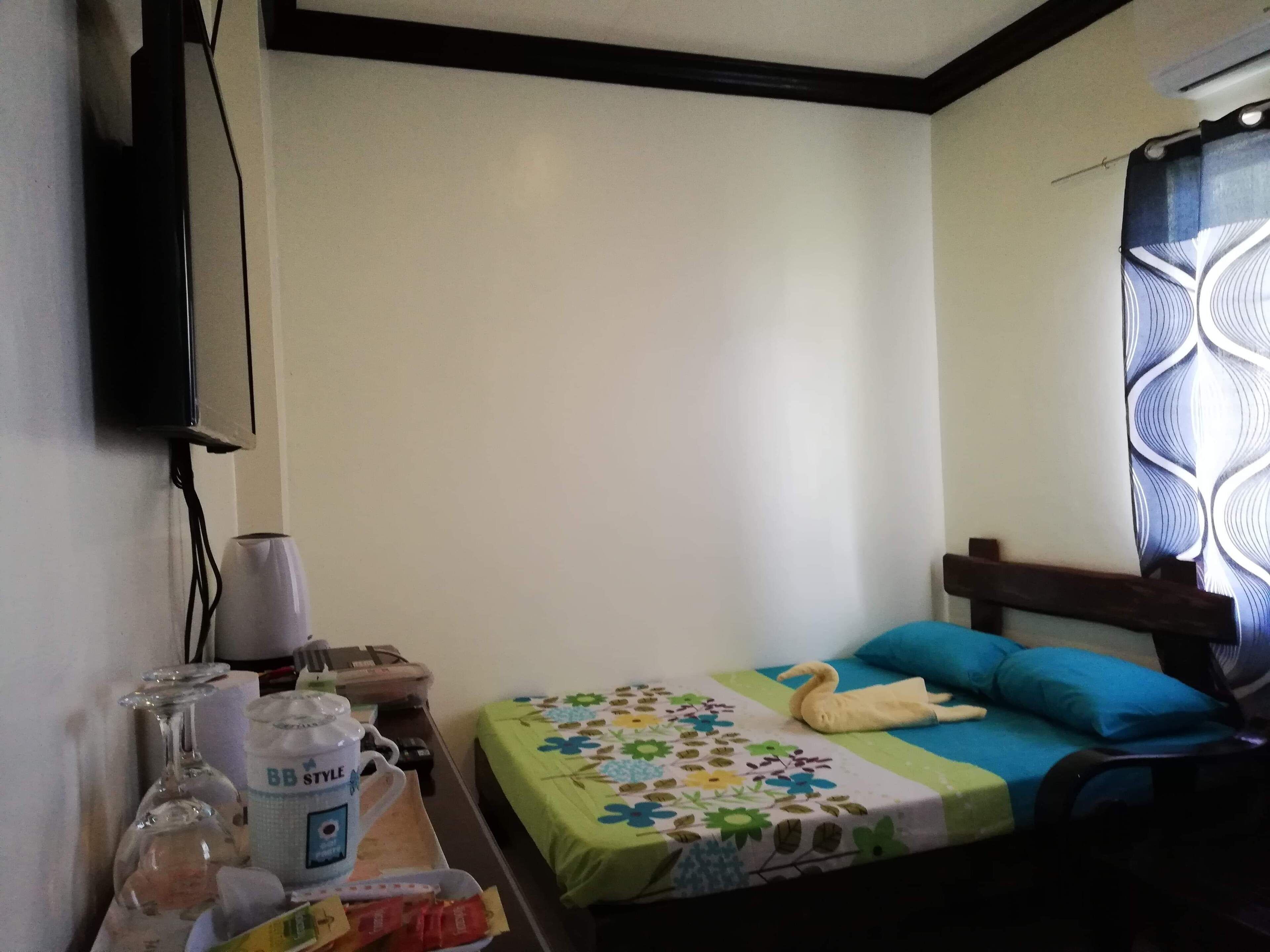 A small, simple room with a bed, a TV mounted on the wall, and a window with a patterned curtain.