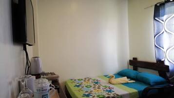 A small, simple room with a bed, a TV mounted on the wall, and a window with a patterned curtain.