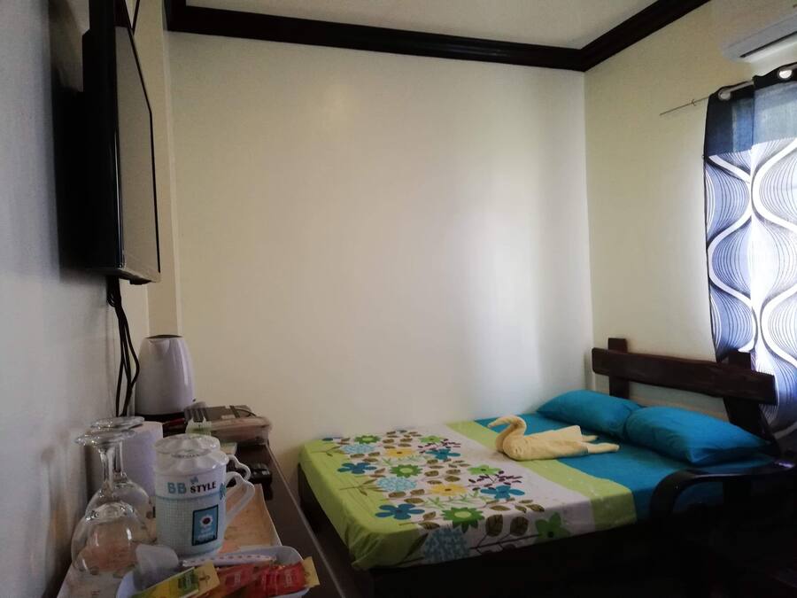 A small, simple room with a bed, a TV mounted on the wall, and a window with a patterned curtain.