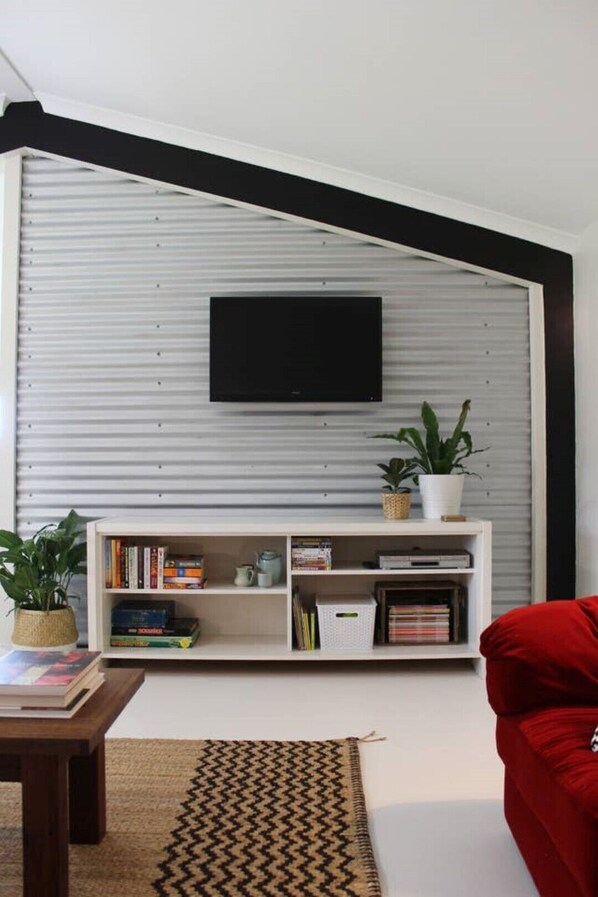 Una sala de estar moderna con un televisor de pantalla plana montado en una pared blanca, una estantería blanca con libros y objetos decorativos, un sofá rojo y una alfombra estampada.