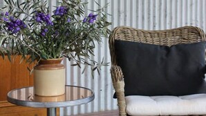 Une chaise en osier avec un coussin noir et une petite table sur laquelle est posé un vase rempli de fleurs violettes.