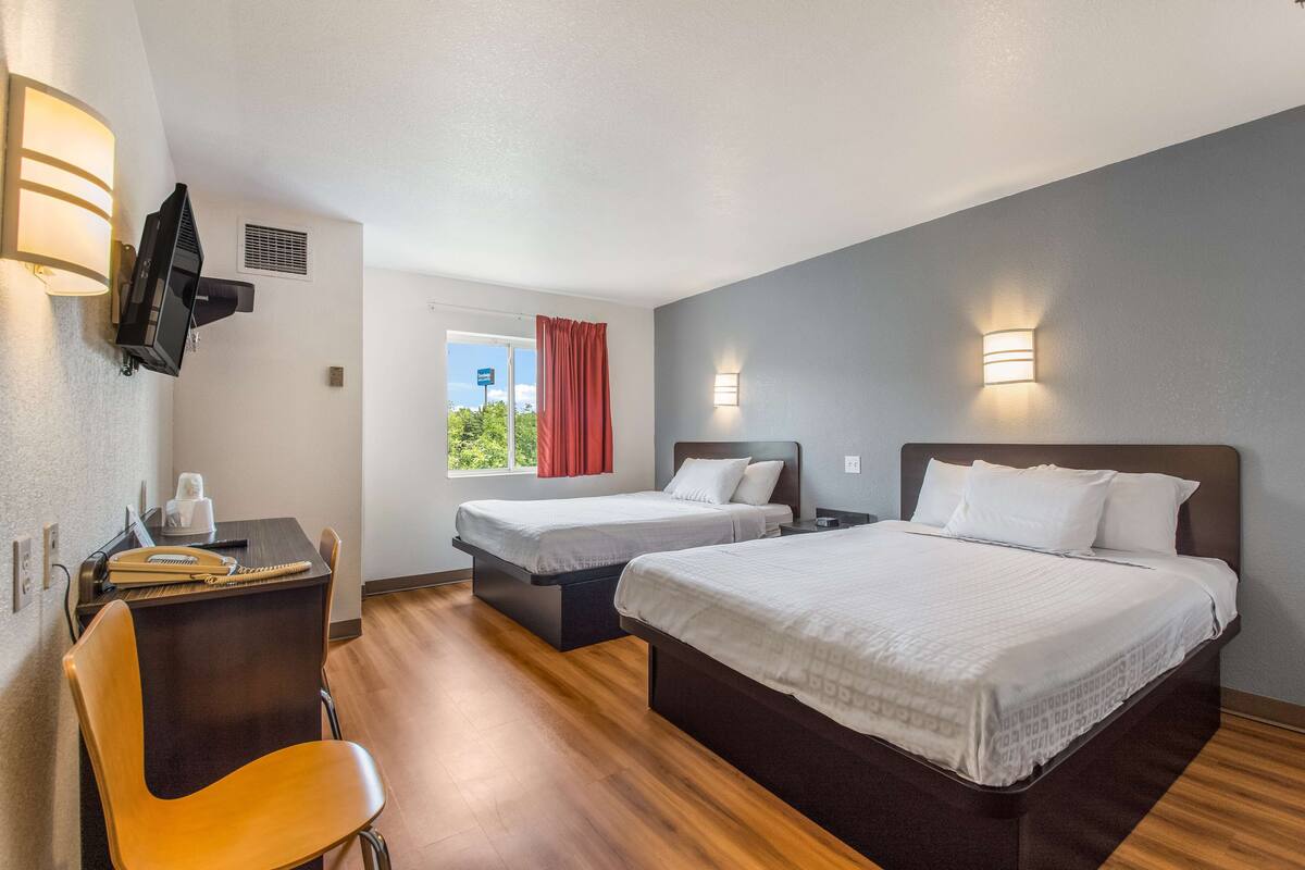 Ein Hotelzimmer mit zwei Betten, einem Schreibtisch mit Stuhl, einem an der Wand befestigten Fernseher und einem Fenster mit rotem Vorhang.