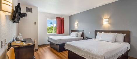 Habitación de hotel con dos camas, un escritorio con una silla, un televisor montado en la pared y una ventana con cortina roja.