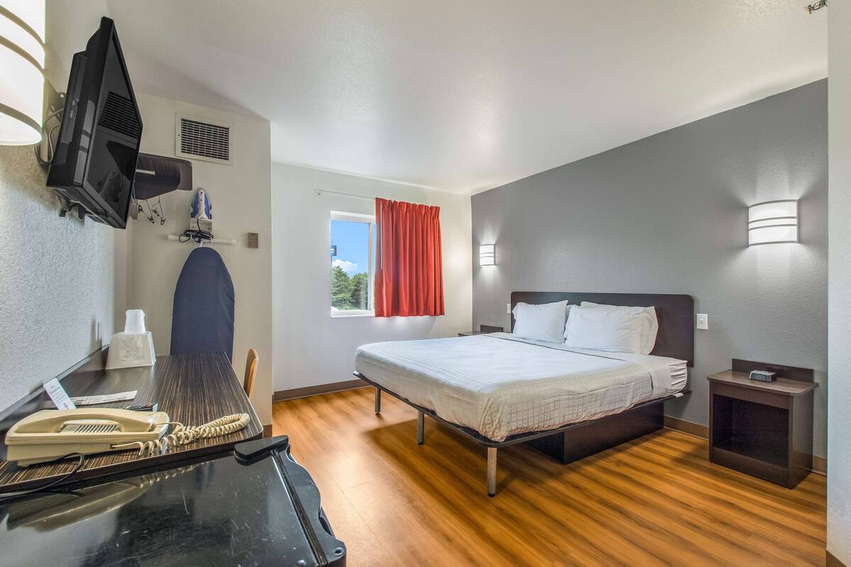Ein Hotelzimmer mit einem Bett, einem Nachttisch, einem Schreibtisch mit Telefon und einem an der Wand befestigten Fernseher.