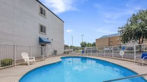 Une piscine entourée d’une clôture blanche, avec une chaise blanche à proximité et un bâtiment visible à l’arrière-plan.