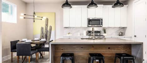 Una cocina moderna con una isla central, taburetes de bar, una zona de comedor con mesa y sillas, e iluminación colgante.