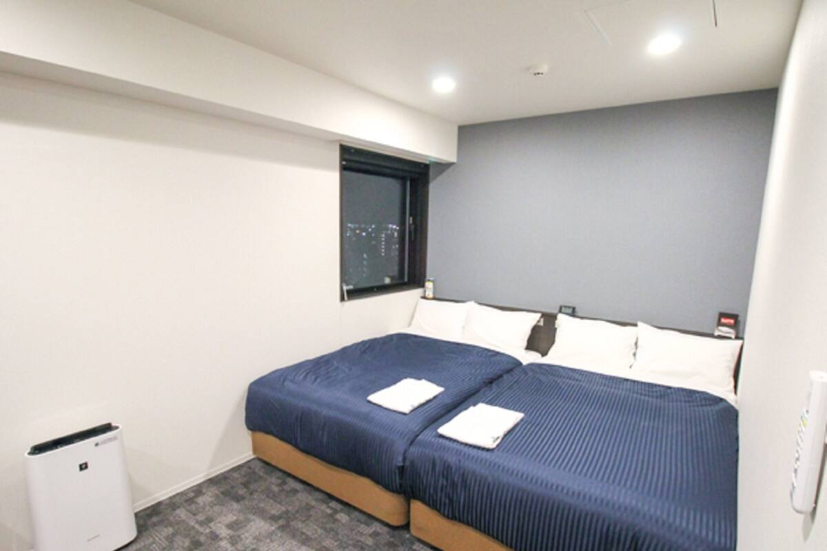 A hotel room with a double bed, a small window, a white trash bin, and a grey carpet.