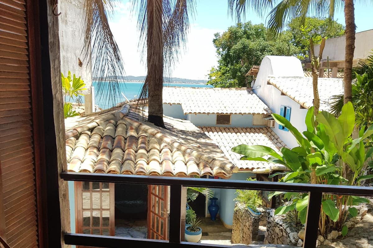 A wooden bench on a balcony overlooking a tiled patio with a view of the sea and palm trees.