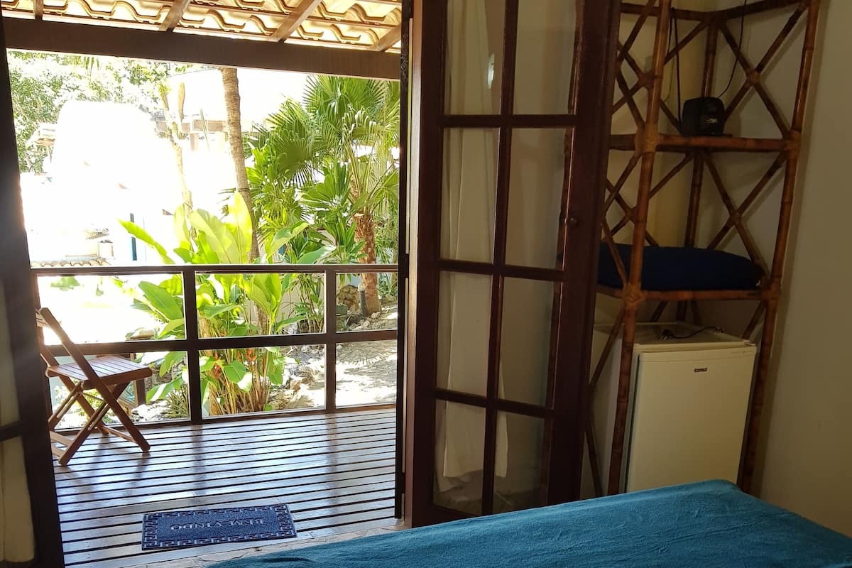 A room with a bed, a wooden chair, a small table, and a view of greenery outside.