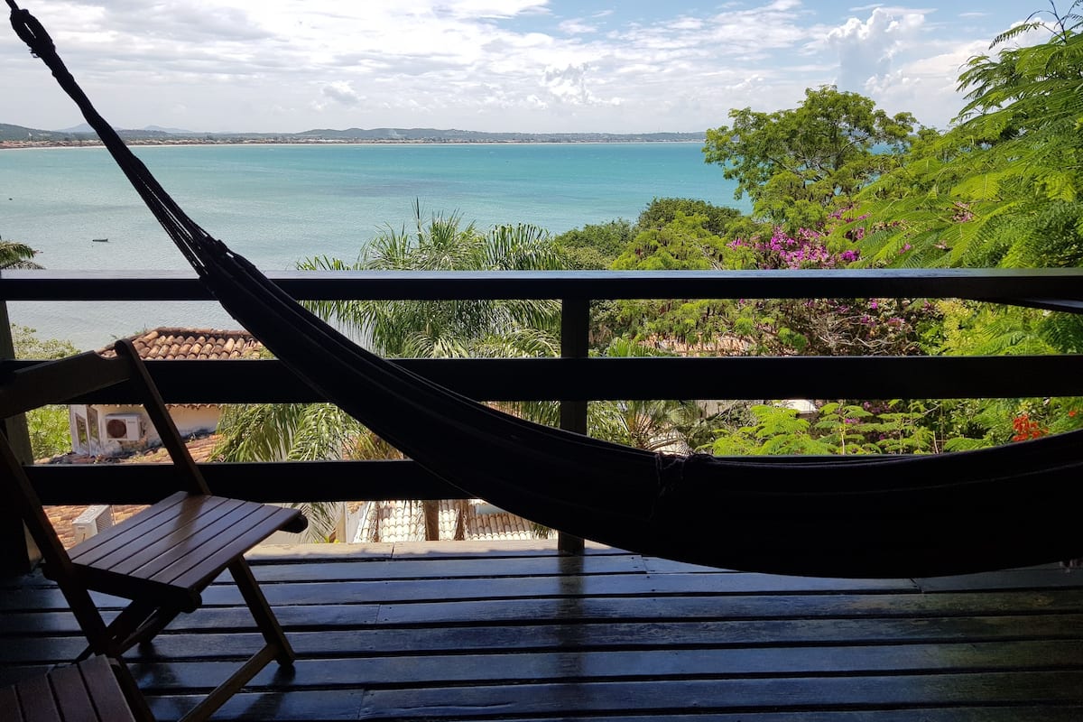 A hammock on a wooden deck overlooking a turquoise sea with a view of distant hills and lush greenery.