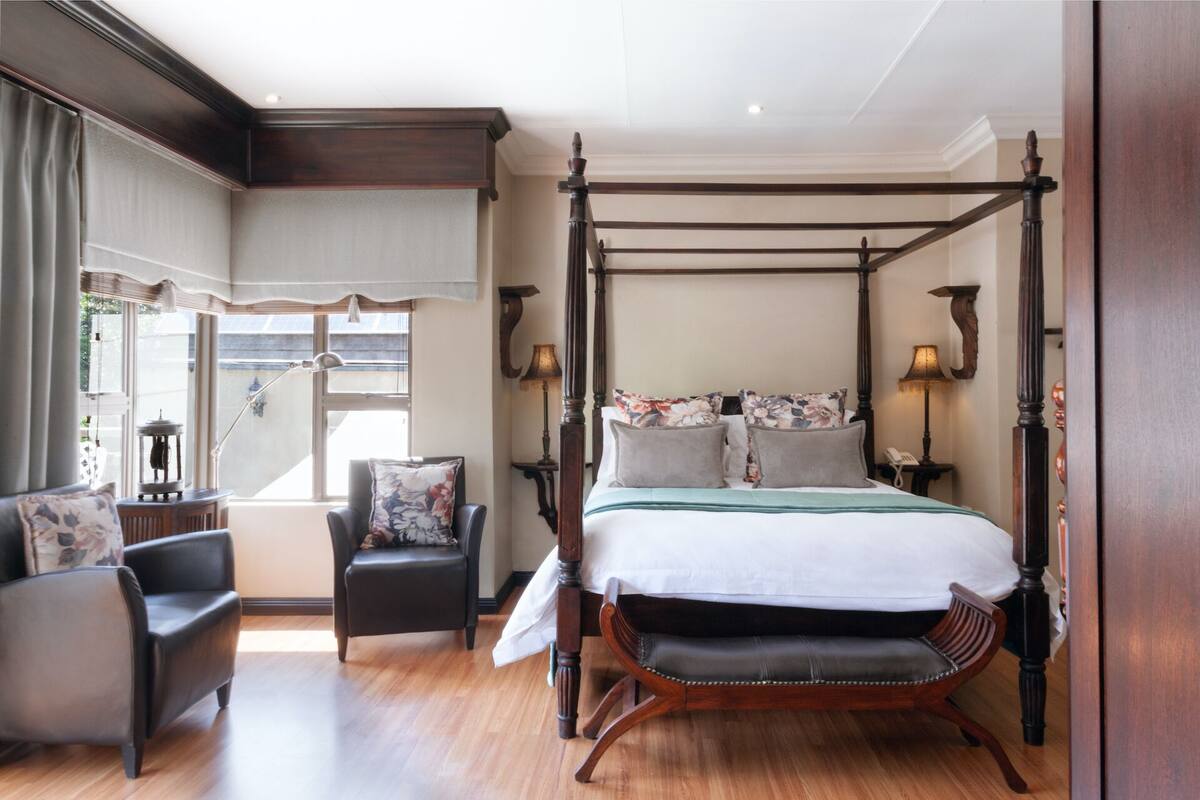 Een hemelbed met bloemenkussens, een houten stoel en een nachtkastje met een lamp.