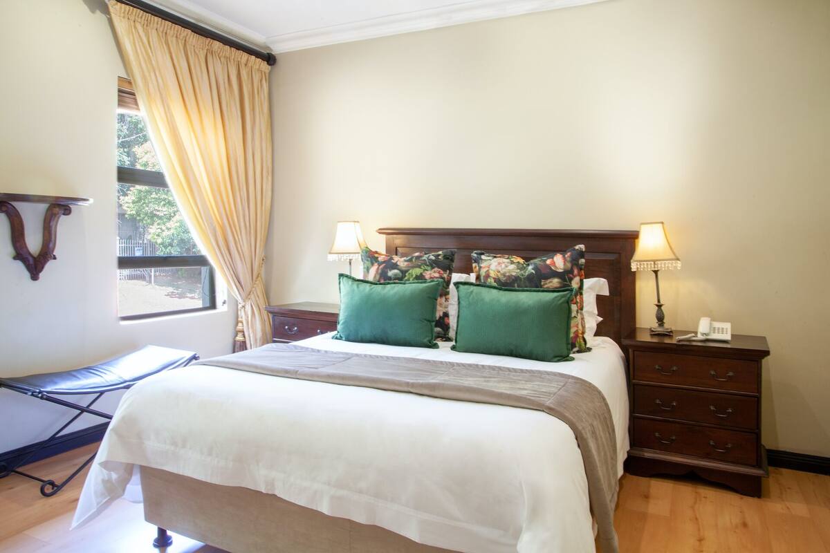 Een net opgemaakt bed met een witte deken en twee groene kussens, een houten nachtkastje met een lamp en een telefoon, en een raam met beige gordijnen.