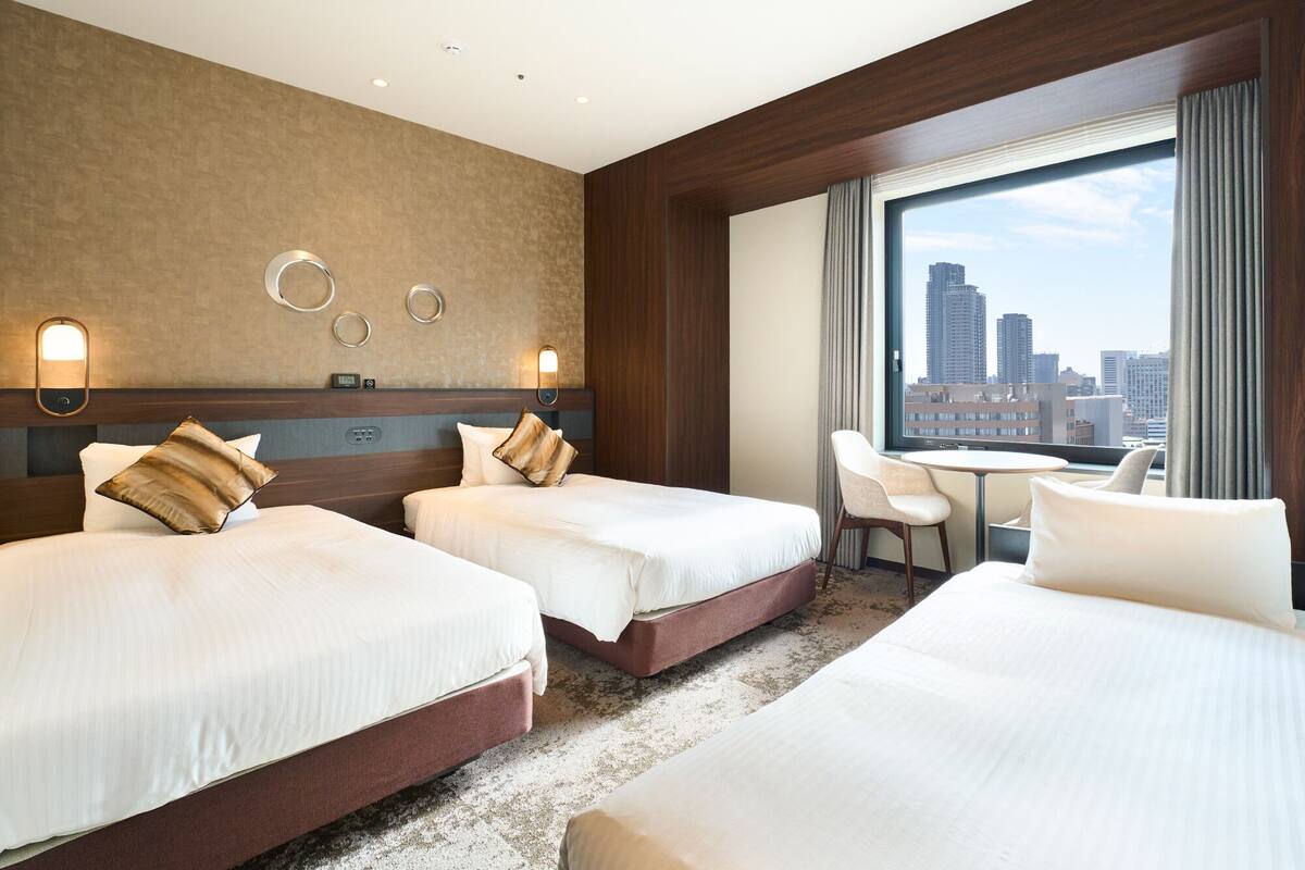 A hotel room with two beds, a desk, and a large window with a city view.