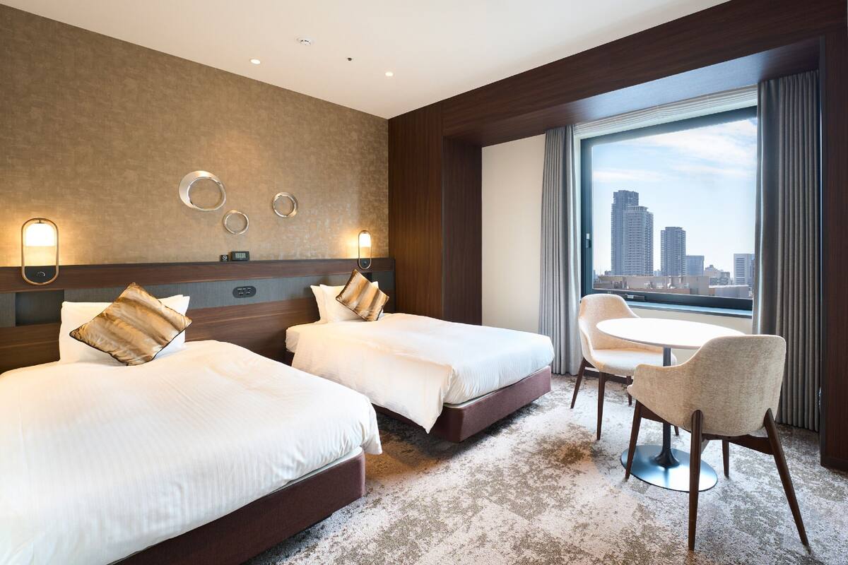 A hotel room with two beds, a small table, and a chair. There are wall sconces and a view of the city.