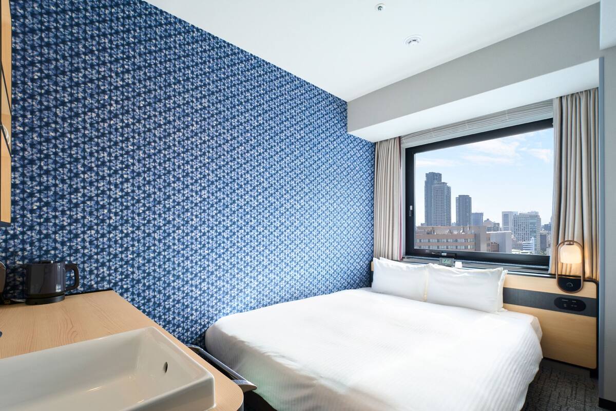 A hotel room with a large bed, a bathroom with a sink, and a city view through the window.