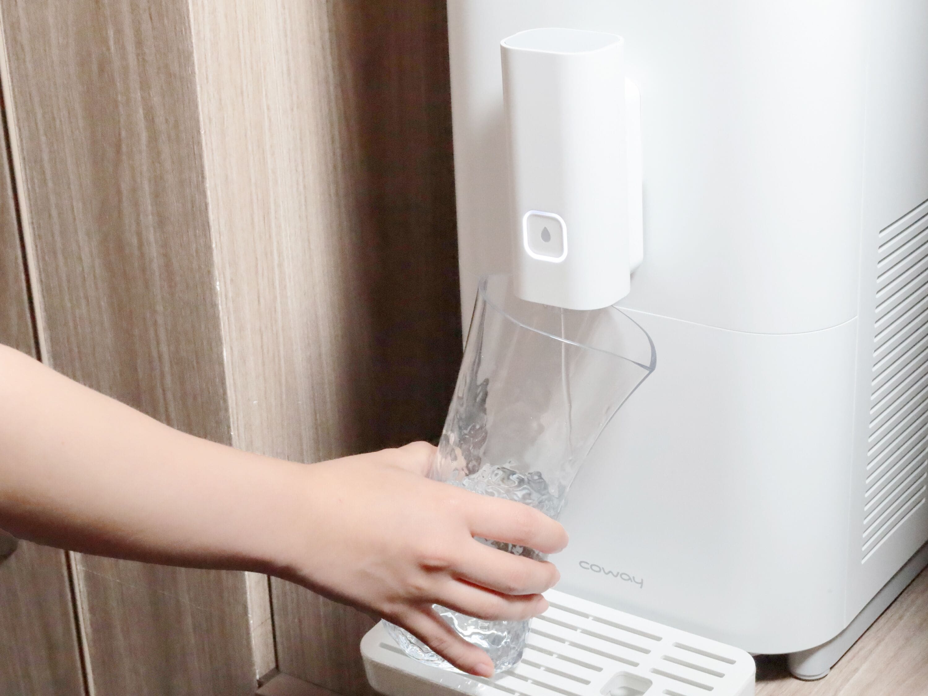 A hand holding a glass of water from a white water dispenser with the brand name 'Coway' visible on the side.