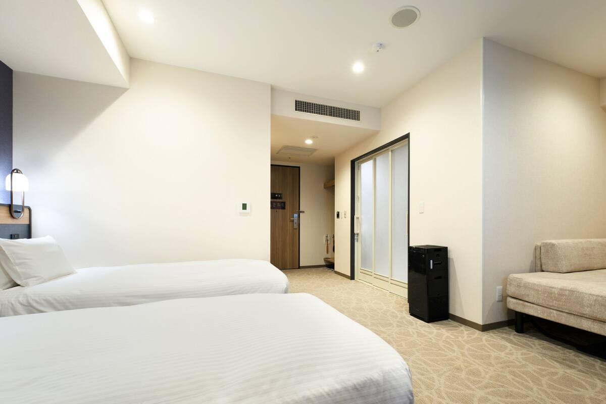 A hotel room with two beds, a sofa, a trash bin, and a door leading to a bathroom.