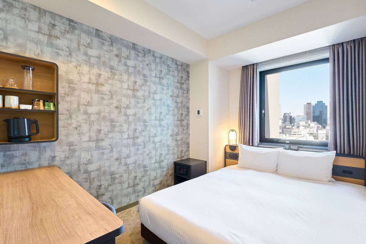 A hotel room with a bed, a wooden desk, a shelf with toiletries, a nightstand, and a window with a city view.