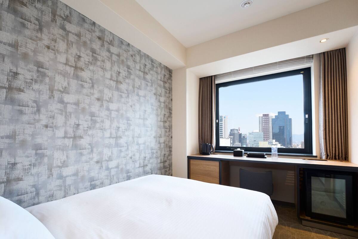 A hotel room with a large bed, a desk with a computer, and a window with a city view.