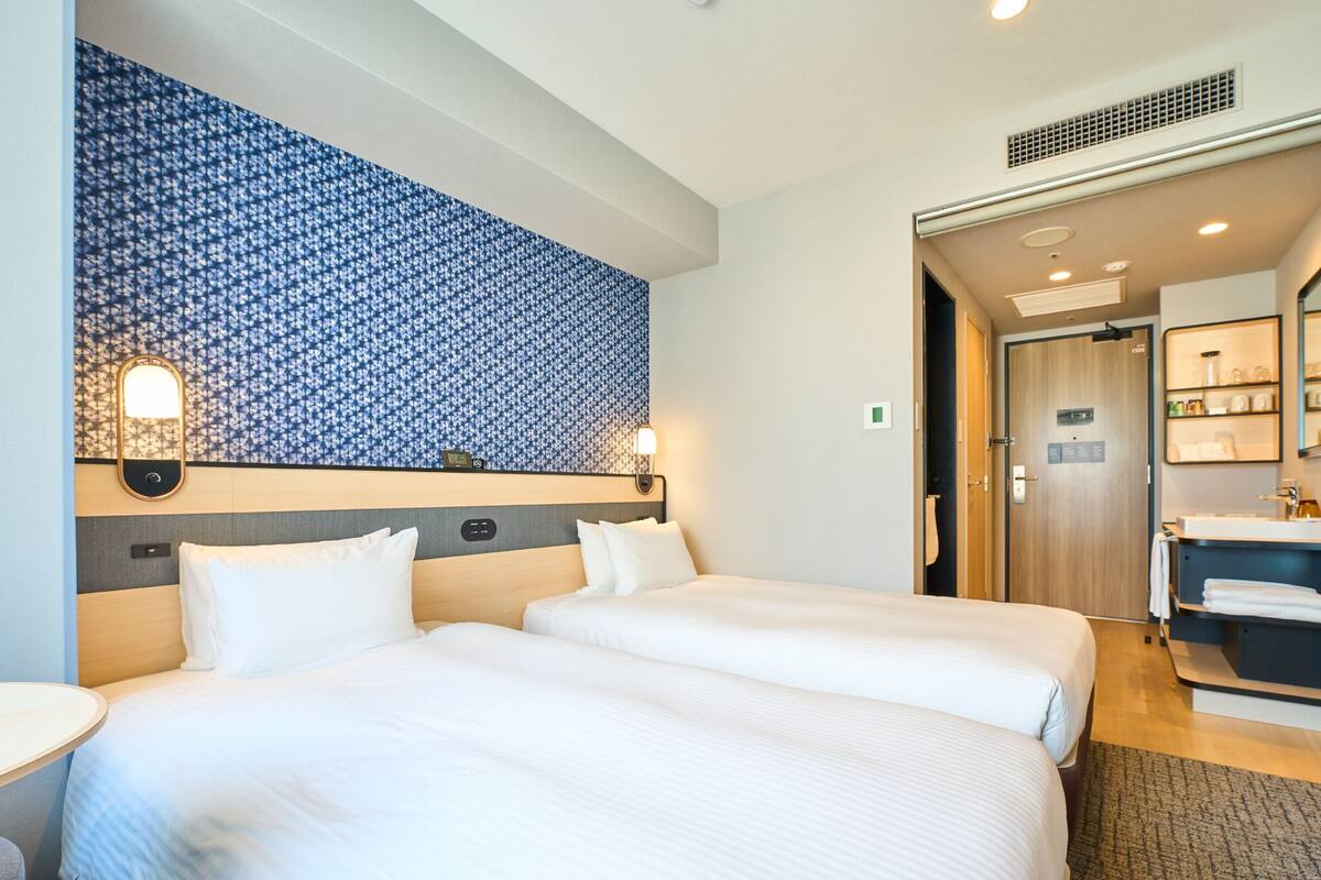 A hotel room with two beds, a bathroom with a sink and mirror, and a patterned wall.
