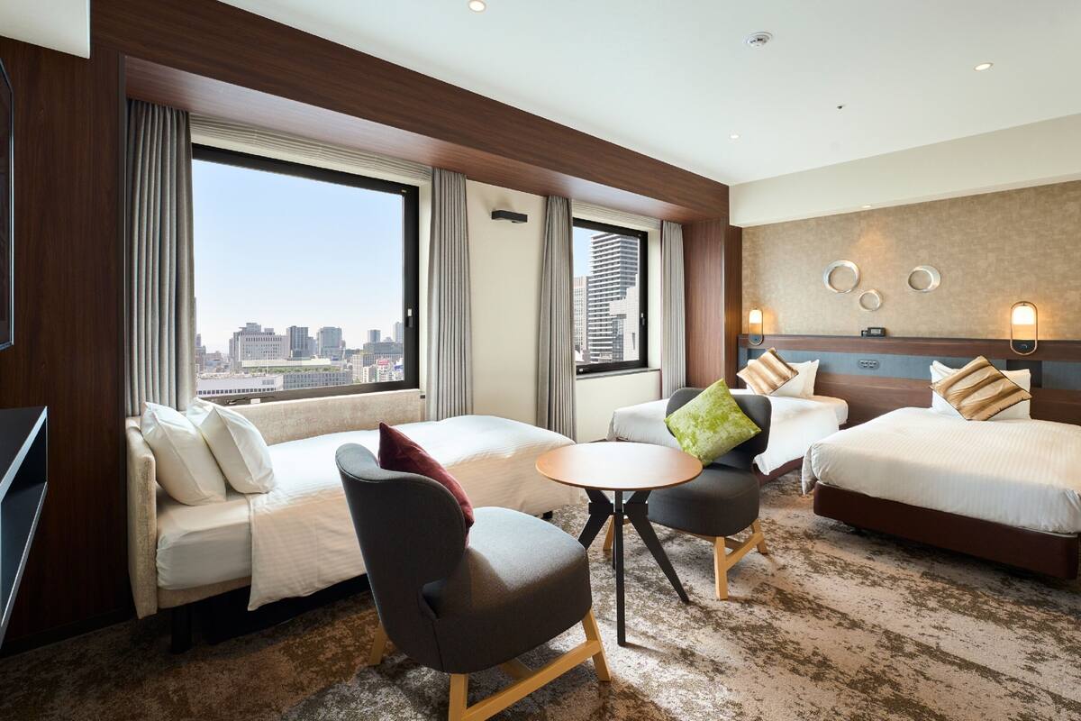 A hotel room with two beds, a sofa, a small table, and a chair. There is a city view through large windows.