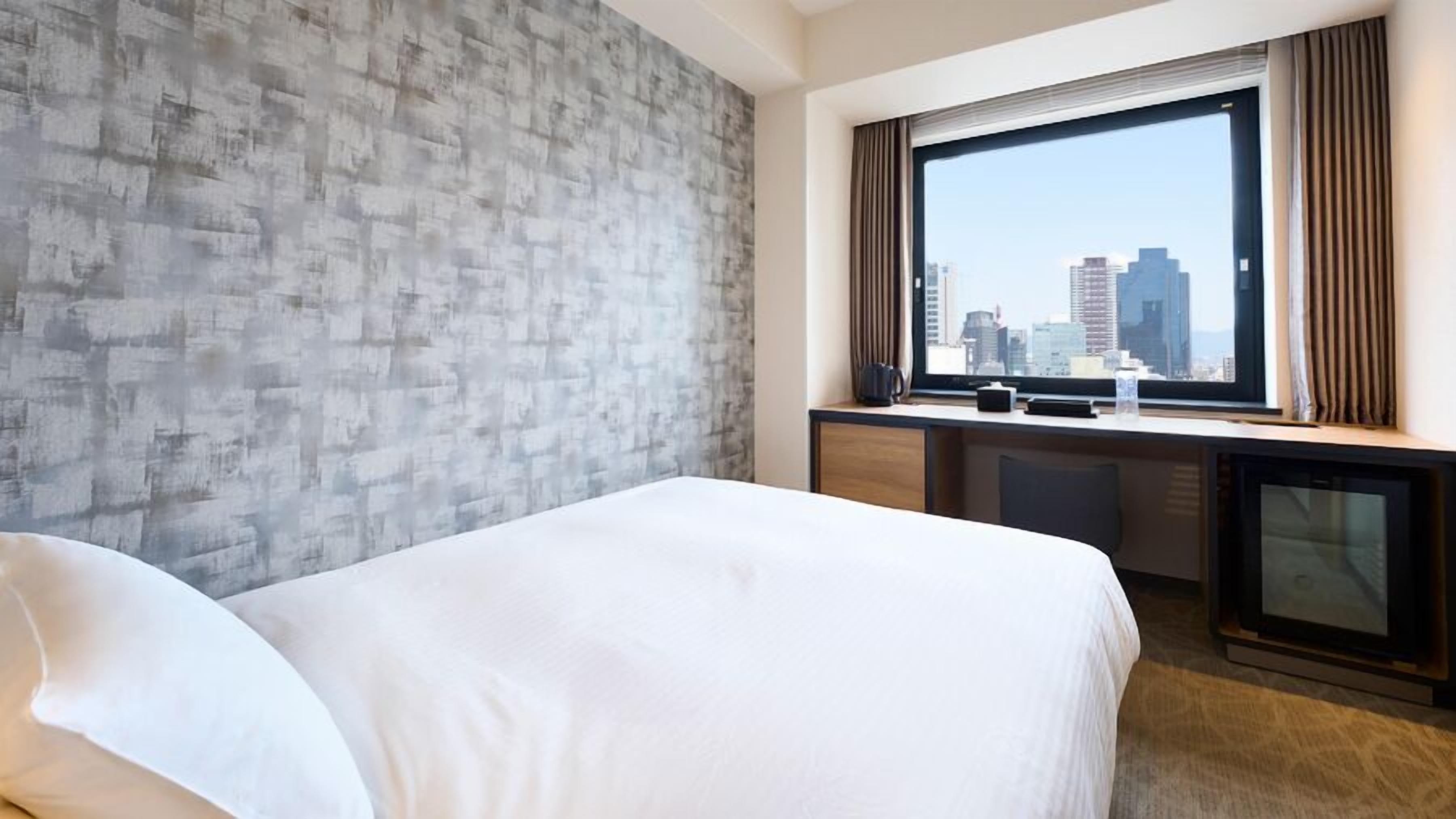 Quarto de hotel com cama, escrivaninha, janela e vista para a cidade, e parede com textura.