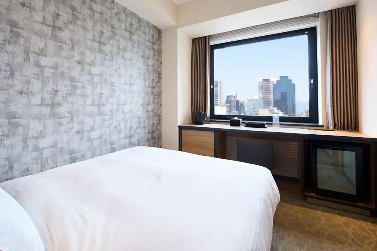 A hotel room with a bed, a desk, a window with a city view, and a textured wall.