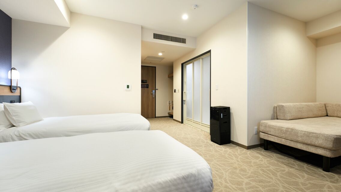 Quarto de hotel com duas camas, um sofá, um cesto de lixo e uma porta deslizante para um banheiro.