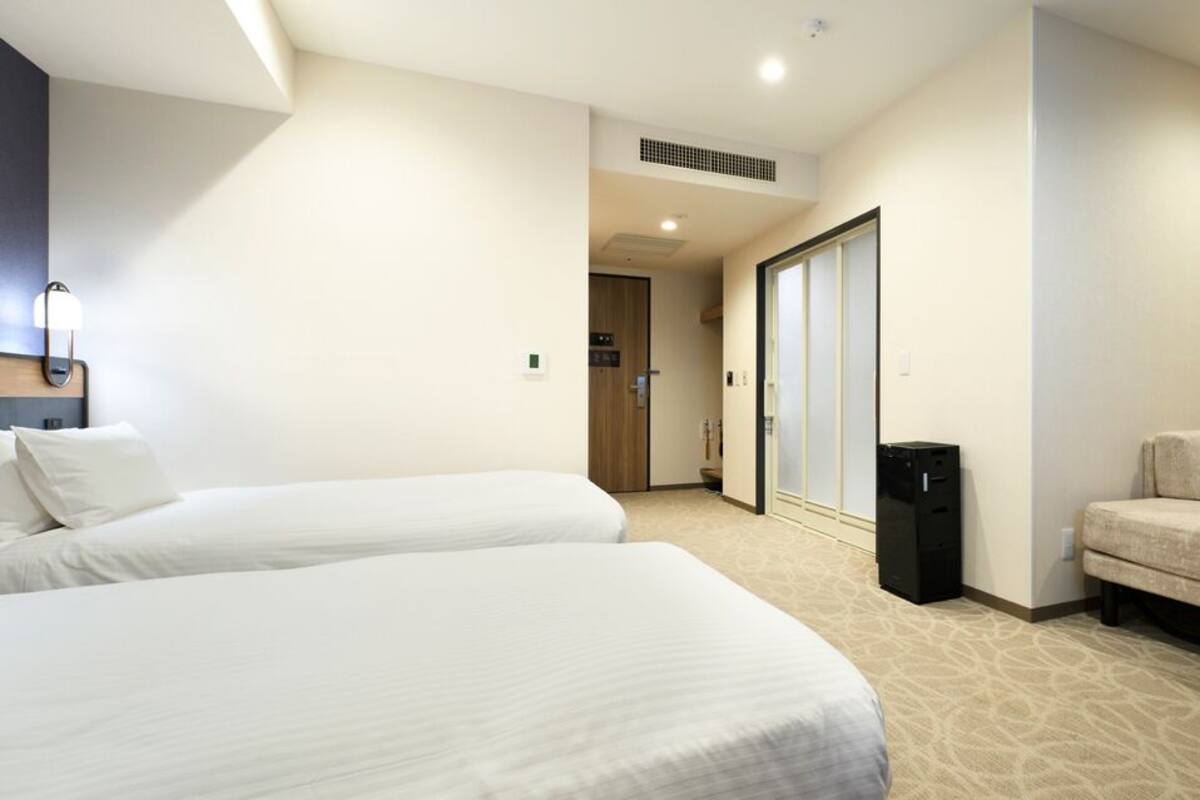 A hotel room with two beds, a sofa, a trash bin, and a sliding door leading to a bathroom.