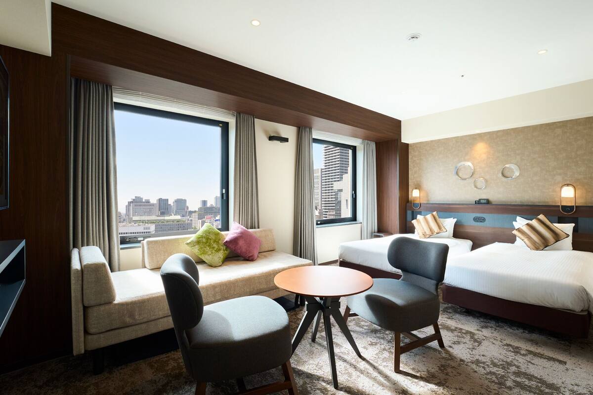 A hotel room with a large window, a sofa, two chairs, a small table, and a bed with multiple pillows.