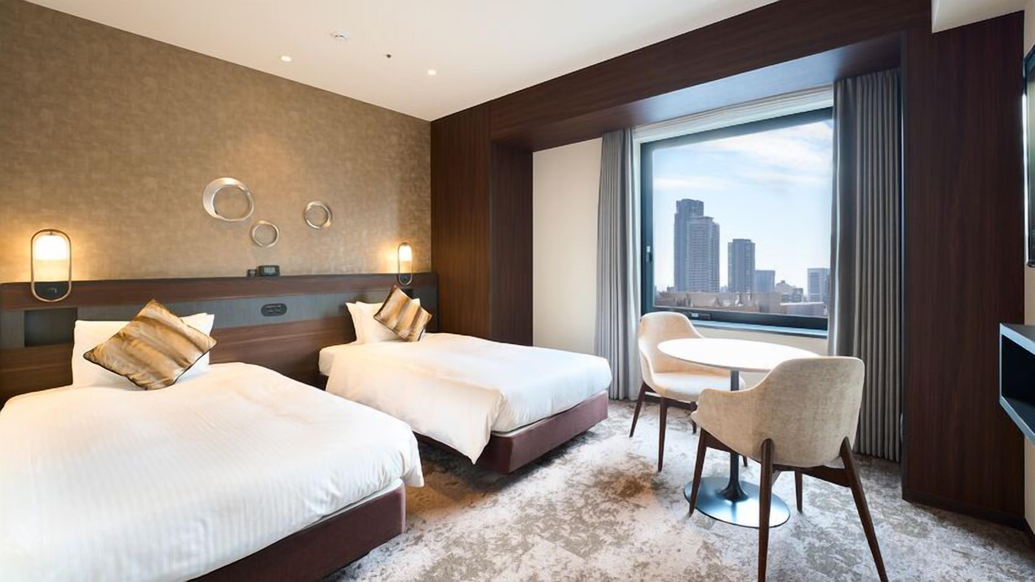 Quarto de hotel com duas camas, uma mesa redonda com cadeiras, uma janela grande com vista para a cidade e iluminação embutida na parede.