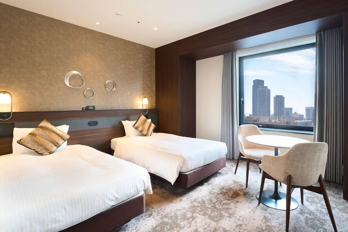 A hotel room with two beds, a round table with chairs, a large window with a city view, and wall-mounted lighting.