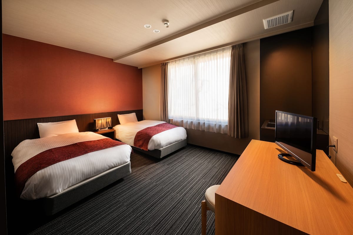 A hotel room with two beds, a desk, a TV, and a window with curtains.