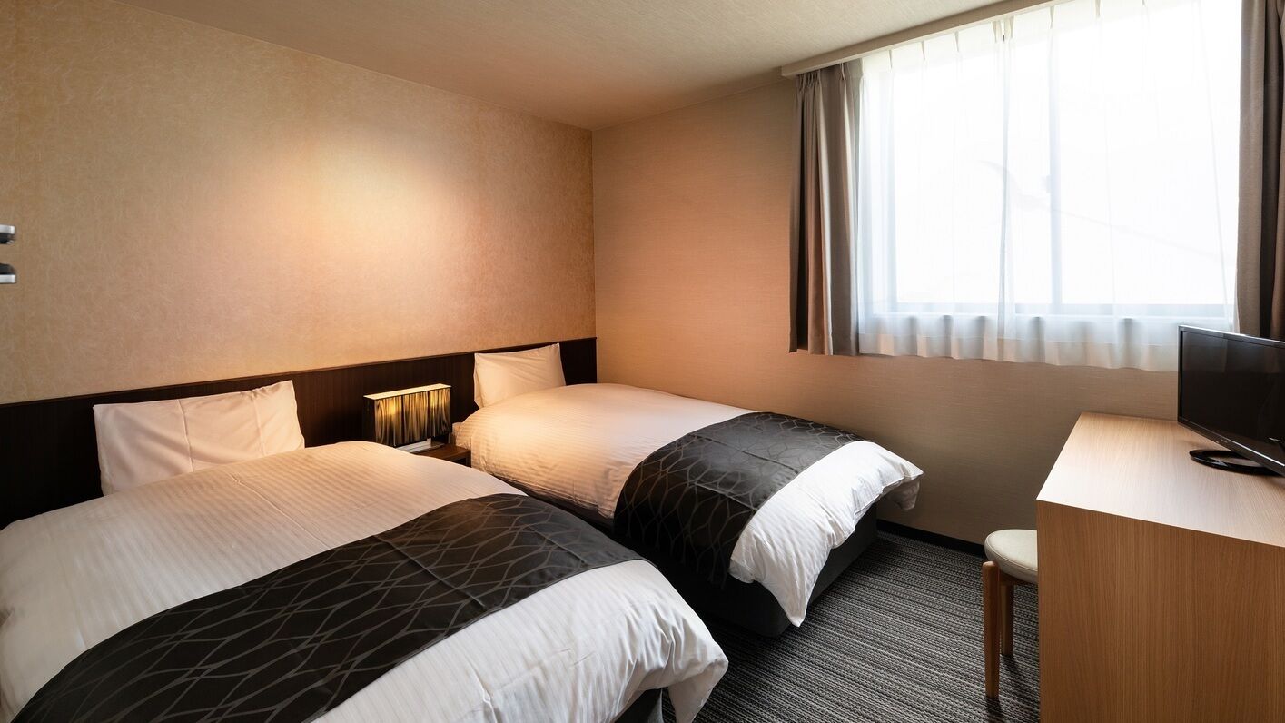 Ein Hotelzimmer mit zwei Betten, einem Schreibtisch, einem Fernseher und einem Fenster mit Vorhängen.