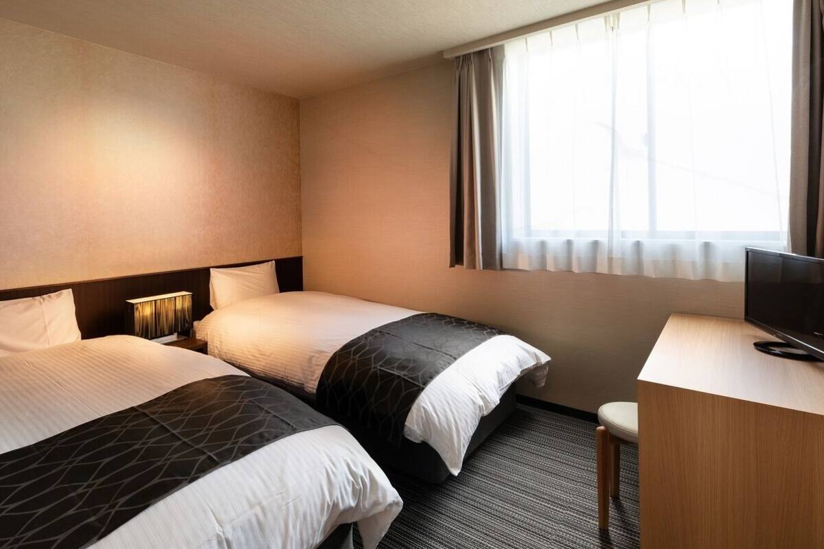 A hotel room with two beds, a desk, a TV, and a window with curtains.