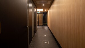 A narrow, dimly lit hallway with wooden paneling on one side and a dark wall on the other. There are two illuminated signs above the door and a small plant near the entrance. - Connect Inn Shichijokamogawa (Kyoto)