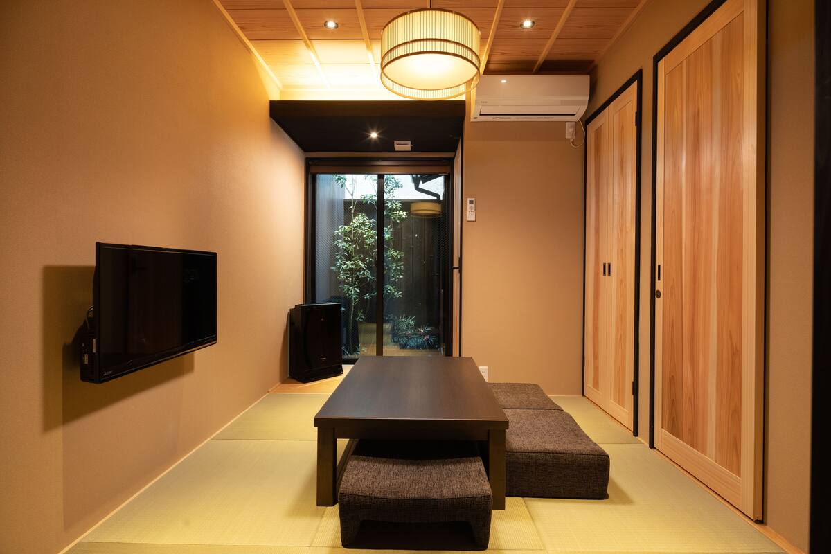 Een traditionele Japanse kamer met tatami vloerbedekking, een lage tafel, kussens, een televisie en schuifdeuren naar een tuin.