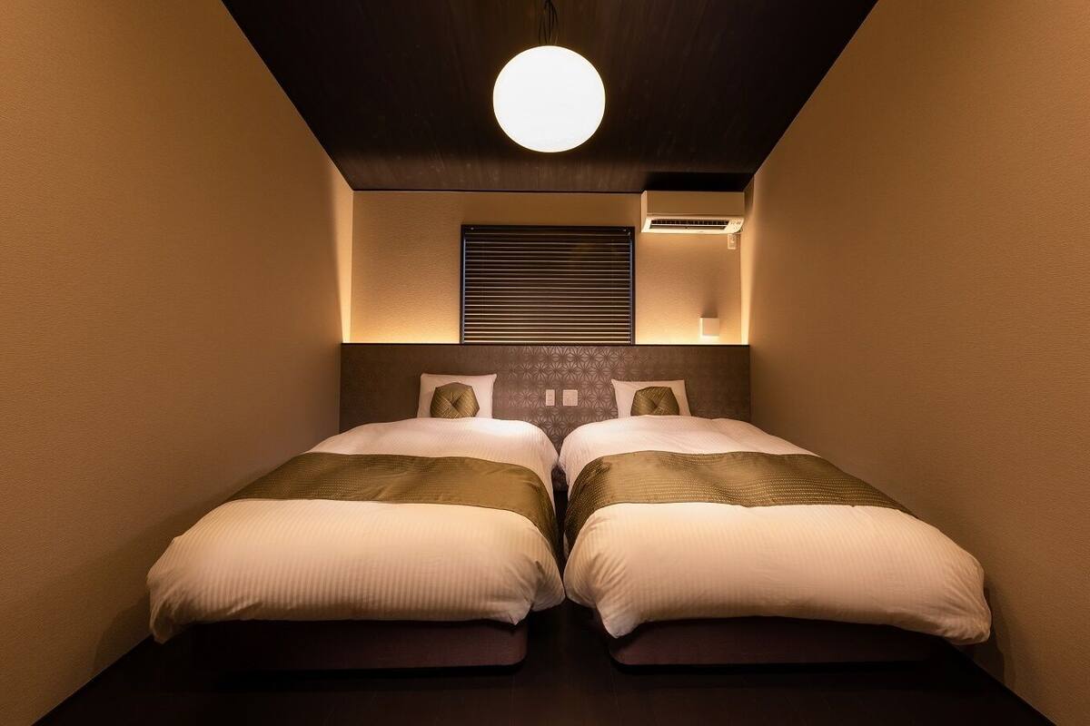 Een kleine hotelkamer met twee bedden, een plafondlamp en een luchtbevoorrachtingsunit aan de muur.