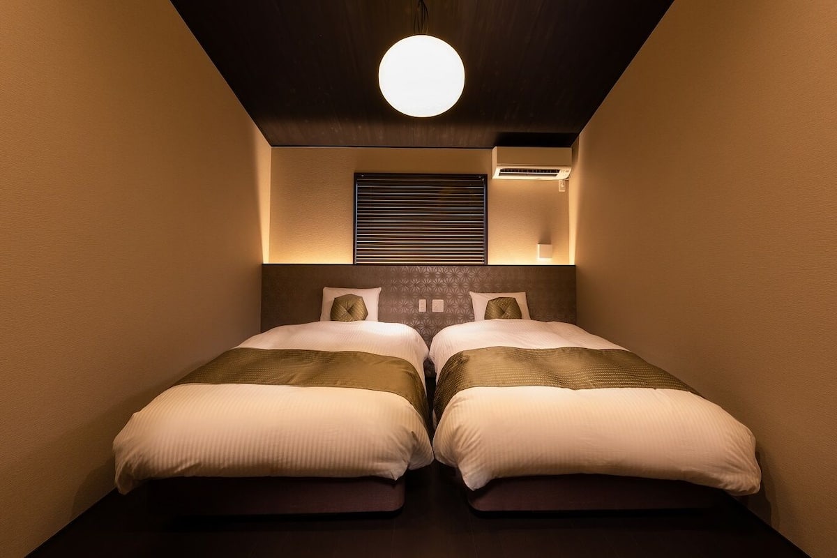 Une chambre d’hôtel étroite, avec deux lits, un luminaire au plafond et un climatiseur fixé au mur.