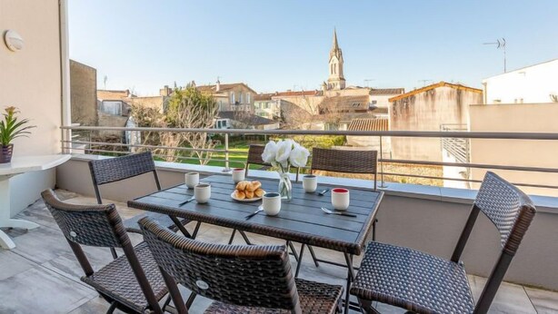 A balcony with wicker furniture, a table with croissants and coffee cups, and a view of buildings and a church steeple. - La Closerie des Tremieres by Interhome (Saint Georges de Didonne)