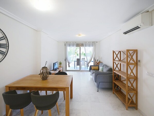 A modern living room with a wooden dining table, two chairs, a grey sofa, a wooden bookshelf, and a large window with curtains.