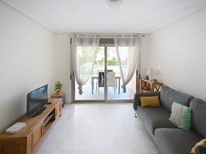 A living room with a flat-screen TV, a grey sofa with cushions, a wooden TV stand, a small table with a lamp, and a view through a glass door to an outdoor area.